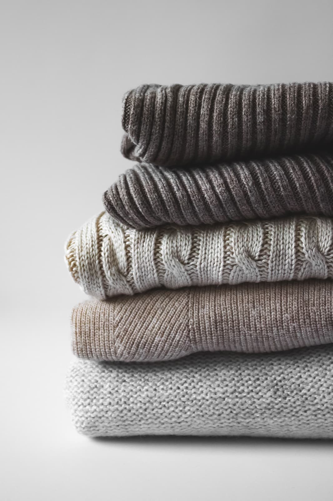A stack of folded knitted garments in undyed cream, grey and brown